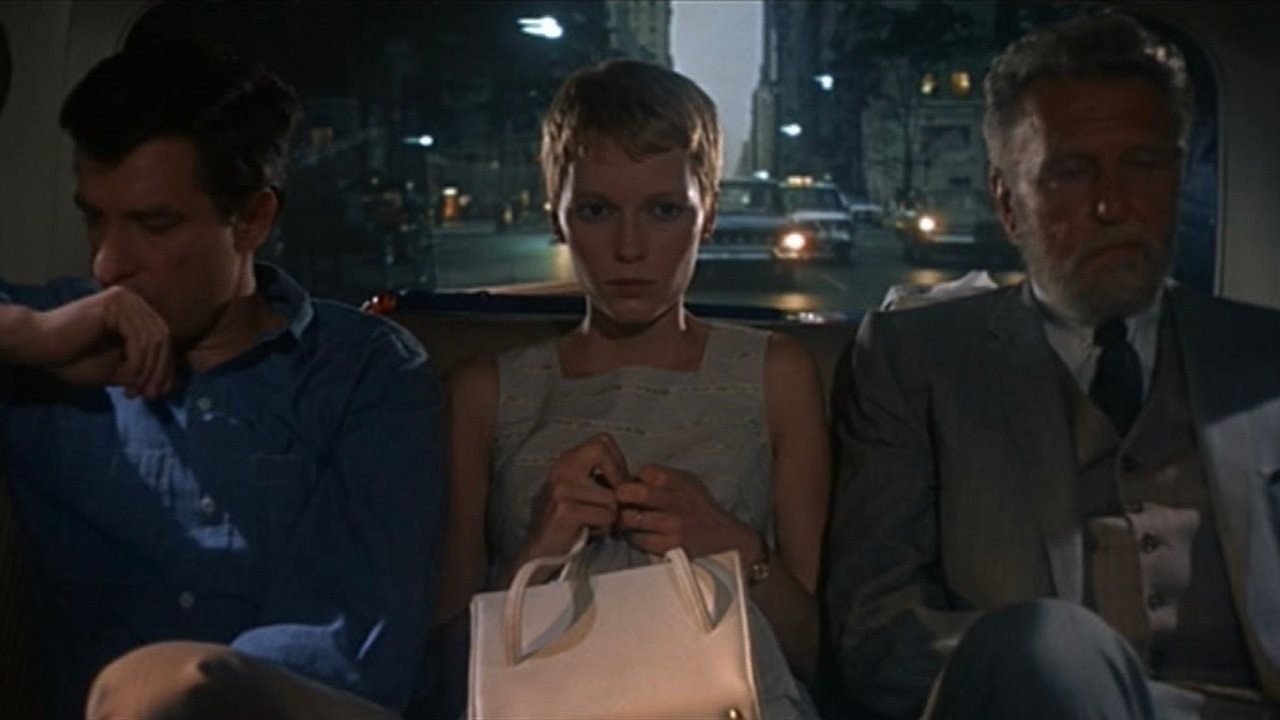 Una joven de pelo corto con bolso blanco, sentada entre dos hombres en un coche de noche, en El bebé de Rosemary (1968)