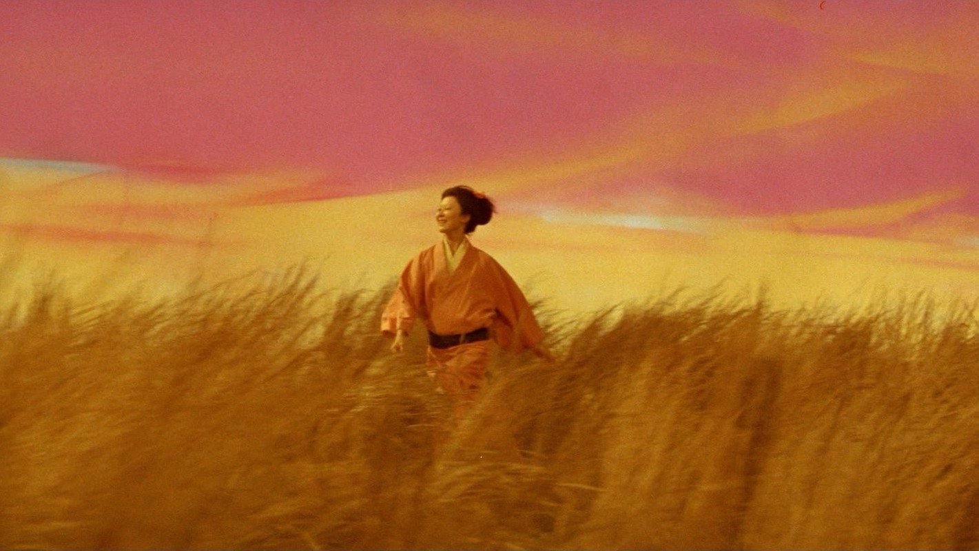 Una mujer con kimono naranja corre entre pastizales bajo un cielo pintado de rosa y amarillo, en Kwaidan (1964)