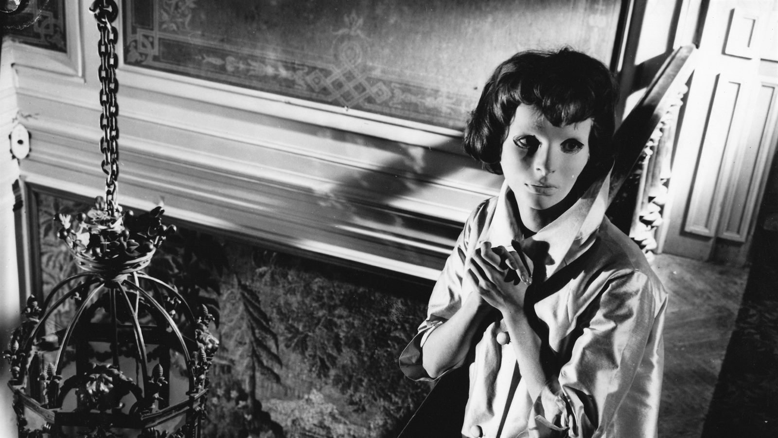 Una joven con máscara blanca inexpresiva y las manos juntas, junto a una lámpara colgante, en Los ojos sin rostro (1960)