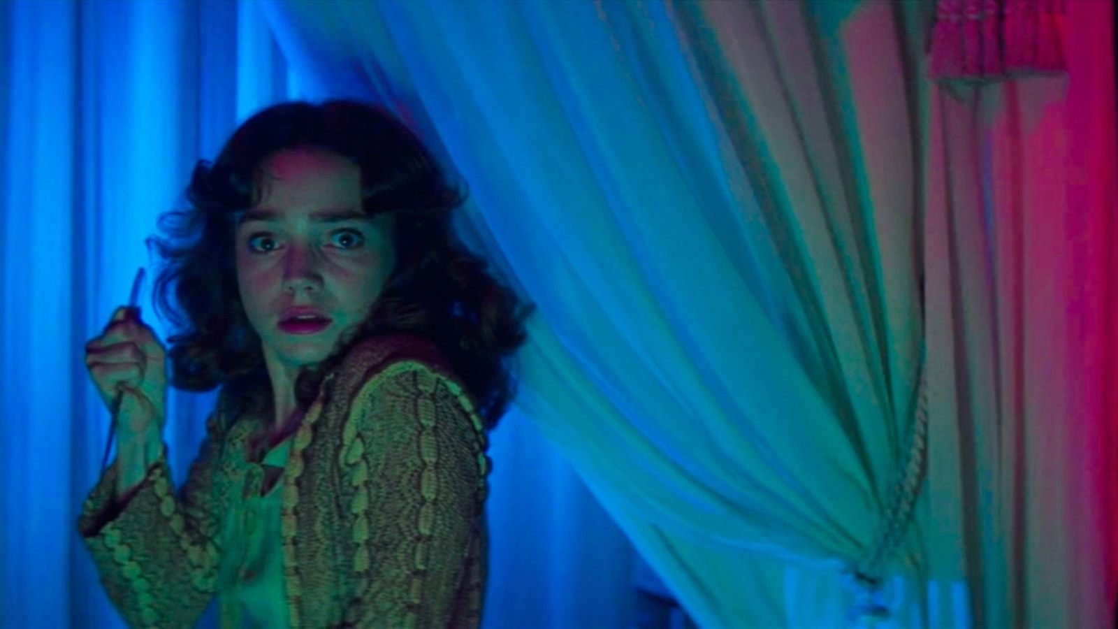 Una joven asustada con un cuchillo se asoma tras una cortina bajo luces azules y rojas, en Suspiria (1977)
