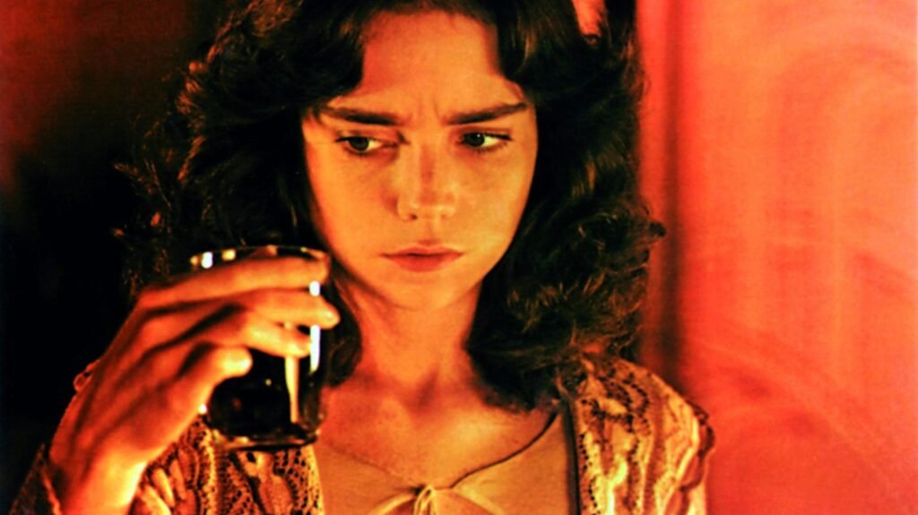 Primer plano de una joven de cabello oscuro que mira un vaso en una habitación bañada en luz roja, en Suspiria (1977)